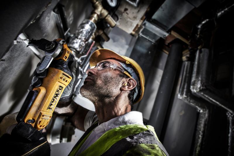 A person wearing safety gear, including a yellow hard hat and high-visibility vest, is using a DEWALT DWE315 multitool to cut through a surface in an industrial setting. The individual's face is obscured.